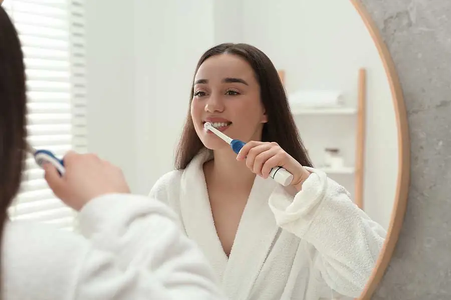 Woman brushing teeth in bathroom mirror.